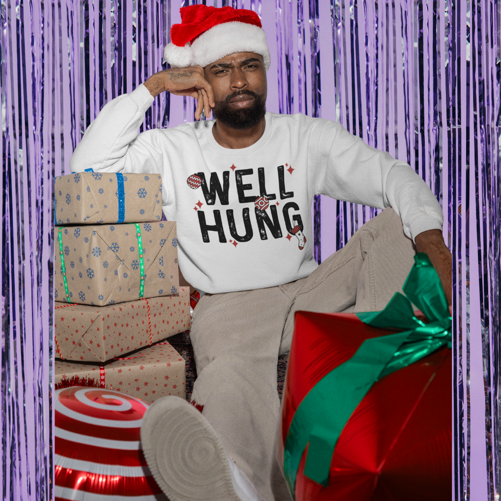 Man wearing a Christmas hat and 'Well Hung' shirt with presents and decorations around him.