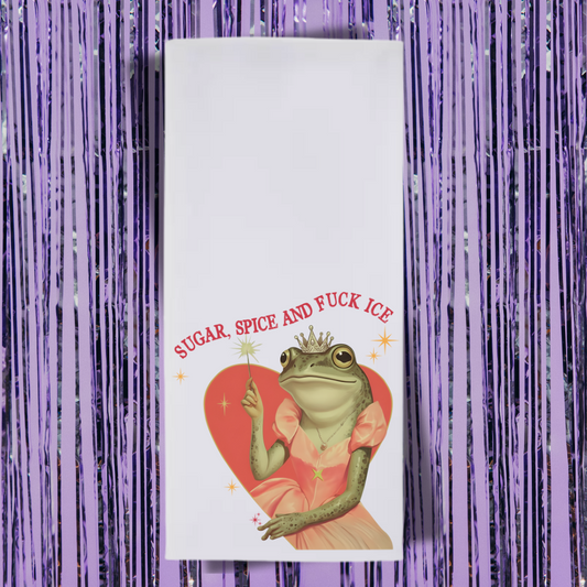 Folded tea towel with a frog wearing a tiara on a heart, text 'SUGAR, SPICE AND FUCK ICE' against a purple tinsel background.